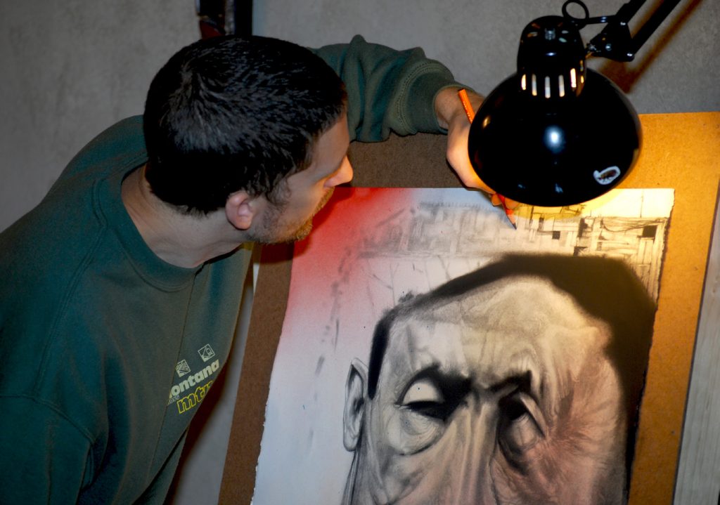 View of the back of the artist as he leans over and works on a charcoal drawing.