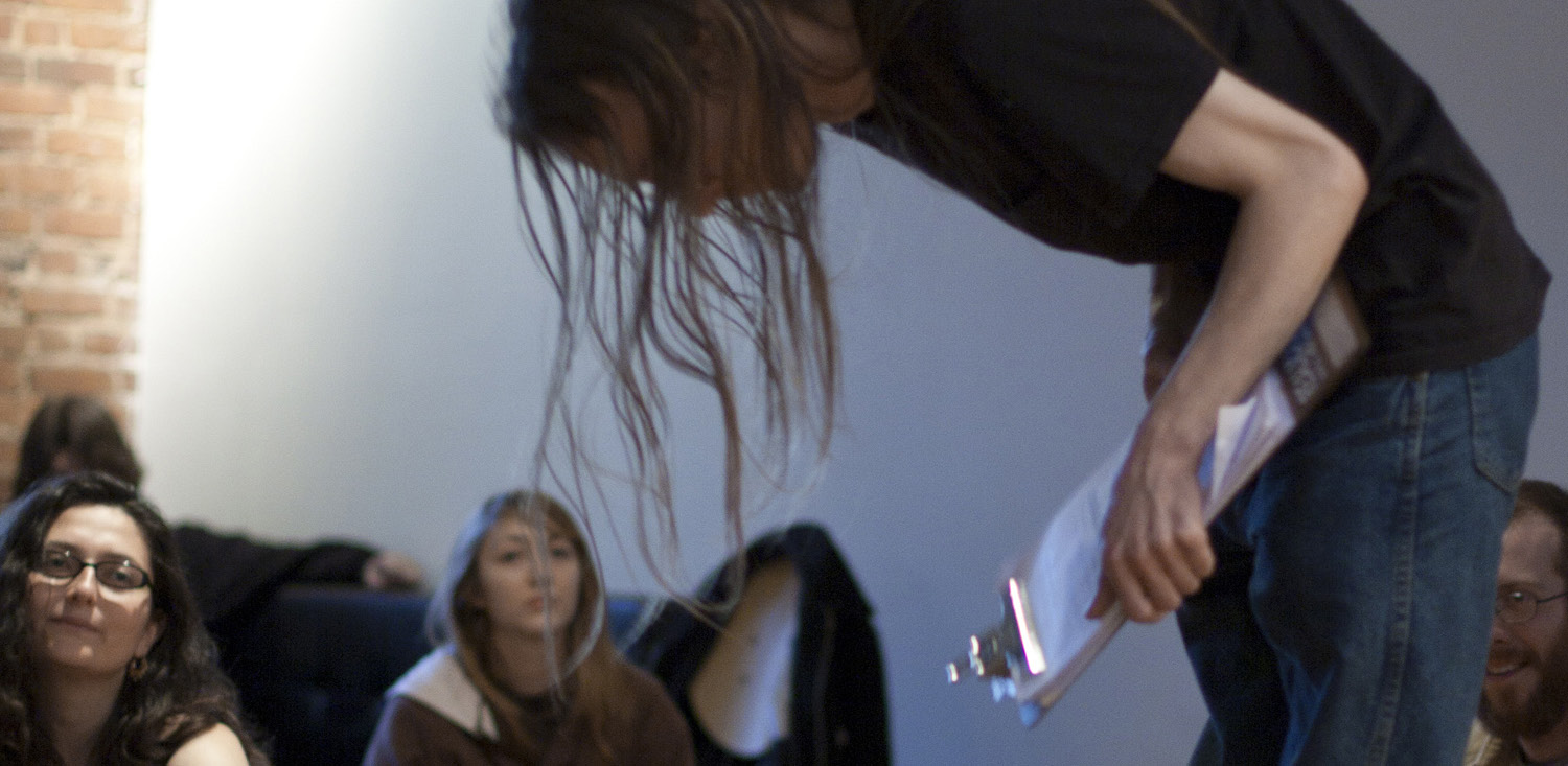 A performance artist with long hair bows in front of a group of seated spectators.